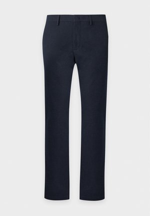 Un homme porte un pull marron, un pantalon bleu marine, des mocassins noirs et un sac à bandoulière sur un fond clair et uni.
