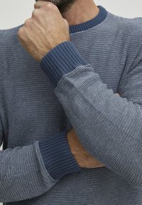 Csíkos kötött pulóver kék árnyalatokban, bordás kerek nyakkal és mandzsettával. Az anyag textúrája puha, váltakozó világos és sötét csíkokkal szőtt.