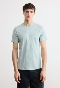 Νεαρός άνδρας με σγουρά μαλλιά, φορώντας ανοιχτό μπλε κοντομάνικη μπλούζα και μαύρο παντελόνι, στέκεται μπροστά σε λευκό φόντο.