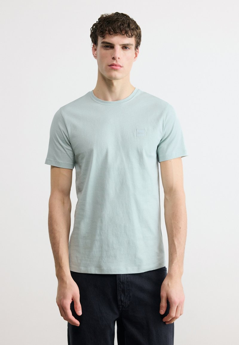 Νεαρός άνδρας με σγουρά μαλλιά, φορώντας ανοιχτό μπλε κοντομάνικη μπλούζα και μαύρο παντελόνι, στέκεται μπροστά σε λευκό φόντο.