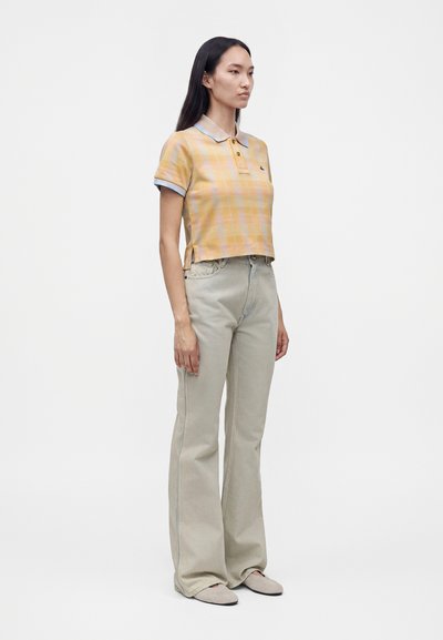Femme debout portant un polo manches courtes à carreaux jaunes, un jean beige taille haute à jambes larges et des chaussures beige clair sur fond blanc uni.
