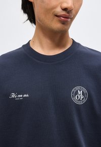 Άνδρας που φοράει σκούρο μπλε φούτερ με λευκό κείμενο "It's on us. Since 1967" στο αριστερό στήθος και στρογγυλό λογότυπο στο δεξί στήθος.