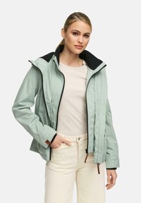 Vrouw in een lichtgroene ritsjack over een beige top en een crèmekleurige broek, staand met één hand in de zak tegen een effen achtergrond.
