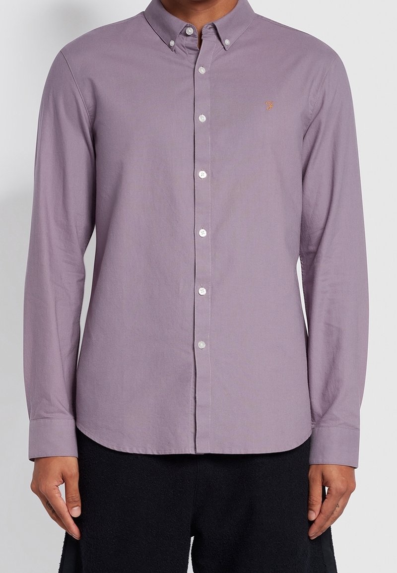 Homme portant une chemise lilas clair à boutons avec col boutonné et logo brodé sur la poitrine gauche.