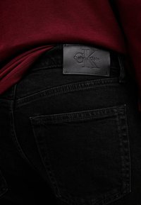 Jeans in denim nero con un tessuto testurizzato, dotati di un'etichetta in pelle Calvin Klein sulla vita e una camicia bordeaux visibile sopra.