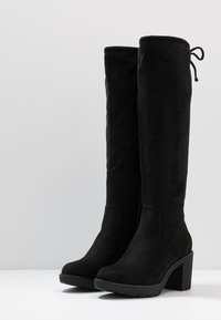 Bottes noires en daim, montantes jusqu'au genou, avec un bout carré, un large talon et un détail de lien à l'arrière. Tige texturée, accents lisses et semelle en caoutchouc robuste.