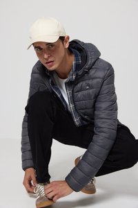 Ung mann iført beige caps, grå puffjakke, rutete skjorte og svarte bukser, sitter på huk og knyter lysebrune sneakers mot en ensfarget bakgrunn.
