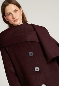 Bordeauxkleurige wollen jas met grote revers, vier zwarte knopen en een elegante uitstraling. Heeft een zachte textuur en een getailleerde pasvorm.