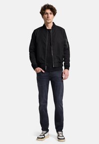 Schwarze Bomberjacke mit Reißverschluss und gerippten Bündchen. Kombiniert mit dunklen Jeans und schwarz-weißen Turnschuhen. Einfaches, strukturiertes Design.