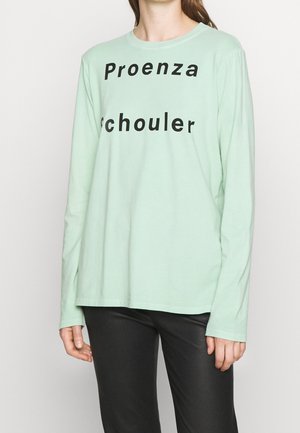 Kobieta ubrana w miętowo-zieloną koszulę z długim rękawem z czarnym nadrukiem "Proenza Schouler", zestawioną z czarnymi spodniami.