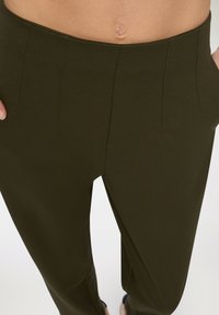 Pantaloni sartoriali verde oliva con una texture liscia, dotati di una vita alta, pieghe frontali e tasche laterali.