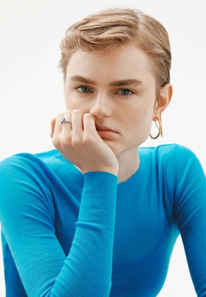 La modelo lleva una blusa de manga larga de color azul brillante. Pendiente de aro dorado y un anillo de plata con una piedra preciosa azul en su mano. Textura de piel suave.