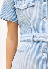 Světle modré džínové šaty s vertikálními bílými pruhy, krátkými rukávy, pasem se stahováním a zapínáním na knoflíky na kapsách. Mají přiléhavý střih a svěží strukturu.