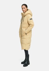 Puffer coat in light beige, knee-length with a hood, zip closure, two pockets, and black drawstrings. Paired with black skinny jeans and ankle boots.