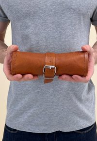 Roulé en cuir marron avec une fermeture à boucle en argent, de forme cylindrique, texture lisse et couture visible, tenu par une personne portant une chemise grise.
