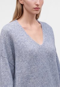 Femme portant un pull en V doux et gris clair à manches longues, montrée des épaules au menton sur un fond clair uni.