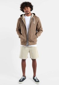 Veste à capuche marron avec fermeture éclair, design bicolore, deux poches avant et intérieur gris. Associée à un short beige et des baskets noires.