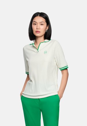 Junge Frau mit glatten schwarzen Haaren trägt ein weißes, geripptes Poloshirt mit grünem Saum und grüne Hose, steht mit den Händen in den Taschen.