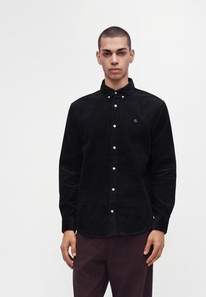 Jeune homme aux cheveux courts porte une chemise noire en velours côtelé boutonnée et un pantalon foncé, debout devant un fond clair uni.