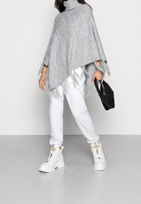 Grå strikket poncho med høj krave, frynset kant, parret med hvide bukser og klodsede hvide sneakers. Komplementerer en sort taske.