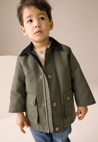 Veste verte avec col noir, fermeture éclair frontale et poches à boutons. Portée sur un pull léger, associée à un jean bleu. Texture lisse.