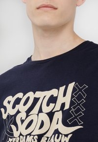 Navyblauwe katoenen t-shirt met grote witte grafische tekst "SCOTCH & SODA" en florale elementen. Rondhals ontwerp, korte mouwen.
