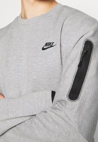Felpa grigia a maniche lunghe con logo Nike nero sul petto e tasca con zip nera sulla manica sinistra. Tessuto morbido e strutturato.