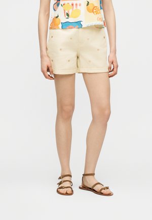 Žena v krémových krátkych nohaviciach s malými srdcovými vzormi, farebnom tričku s ovocným potlačou a hnedých plochých sandáloch so remienkami.