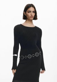 Maglione nero a maglia con maniche lunghe, caratterizzato da una trama a coste e un bracciale argentato. Messo in risalto da una cintura circolare con dettagli borchiati.