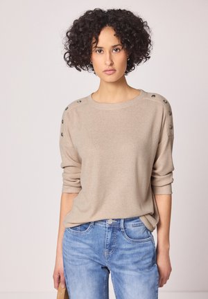 Mujer con cabello rizado vistiendo un suéter beige con botones en los hombros y jeans azules, de pie contra un fondo claro y sencillo.