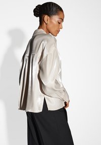 Blusa di satin beige chiaro con design ampio e drappeggiato, maniche lunghe e fessure laterali. Abbinata a pantaloni neri, di colore uniforme.