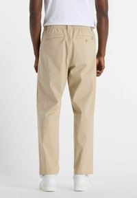 Beige trousers with an elastic waistband, two back pockets, and a relaxed fit; paired with white sneakers. Smooth fabric texture.