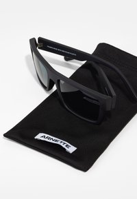 Ochelari de soare negri cu finisaj mat, realizați din plastic reciclat. Îi însoțește un săculeț negru cu eticheta "ARNETTE".