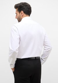 Witte langekost shirt met een klassieke kraag, gladde textuur en knopenmanchetten, gecombineerd met zwarte op maat gemaakte broek, gezien van de achterkant.