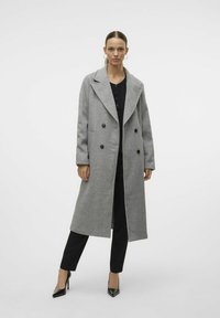 Cappotto grigio in una miscela di lana con design doppiopetto, ampi revers e bottoni neri, indossato sopra un outfit nero e abbinato a tacchi neri.