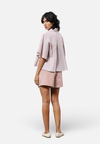 Helle rosa plissierte Bluse mit Glockenärmeln, kombiniert mit rot-weiß gestreiften Shorts. Getragen mit geflochtenen Slippern.