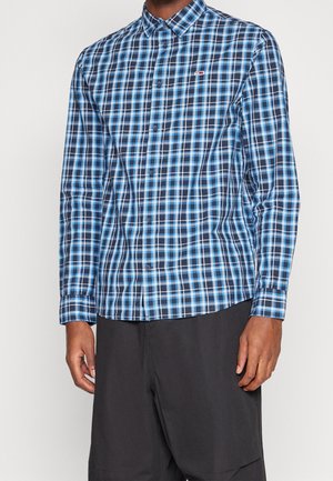 Hombre con camisa de manga larga de cuadros azul y blanco y pantalones negros sobre un fondo liso.