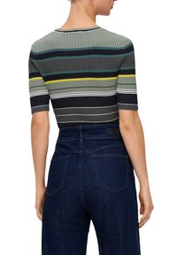 Persoon met een geribbelde, korte mouwtop met horizontale strepen in groen, geel, wit en zwart, gecombineerd met high-waisted donkerblauwe jeans.