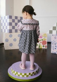 Malé dieťa v kvetinových šatách stojí na kruhovej platforme, obklopené farebnými vzorovanými krabicami v dobre osvetlenom priestore.
