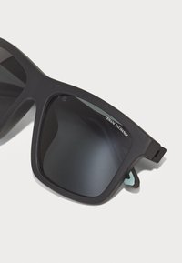 Ochelari de soare din plastic negru, cu rame ușor pătrate și rotunjite, lentile de culoare gri închis și un detaliu subtil al logo-ului pe latură.