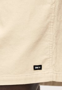 Μπεζ σορτς από βελούδο κοτλέ με οριζόντια ραβδώσεις, ίσια γραμμή και μαύρη ετικέτα με τη λέξη "OBEY" στο κάτω μέρος.