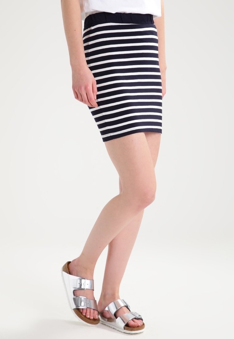 Gonna a righe blu navy e bianche, lunghezza ginocchio con vita elastica. Abbinata a sandali argento con due fibbie su sfondo chiaro.