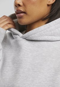 Persona regulē gaiši pelēkā džempera kapuci, fokusējoties uz apakšējo sejas daļu, roku un auduma tekstūru.