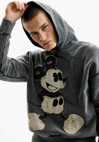 Fiatal férfi rövid hajjal, kopott szürke kapucnis pulóverben, amelyen egy sérült Mickey egér folt látható, miközben az egyik oldalát felemeli a kapucninak.