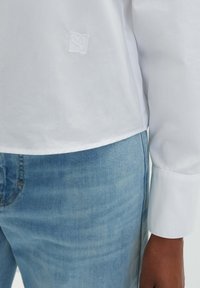 White long-sleeve shirt with embroidered logo, paired with light blue denim jeans. The shirt has a smooth texture and a straight hemline.