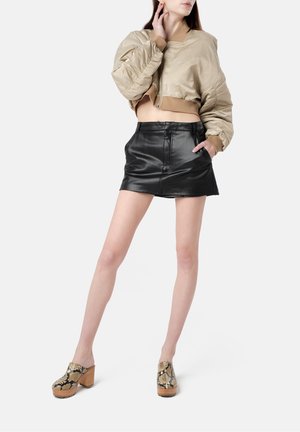 Giacca bomber beige corta, mini gonna in pelle nera e zoccoli in pelle di serpente con tacchi in legno. Materiali morbidi e design aderente.