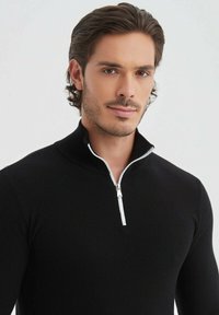 Sweatshirt noir zippé avec col montant, texture côtelée et fermeture éclair blanche contrastante. Coupe ajustée avec une finition lisse.