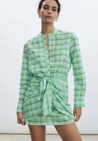 Abito camicia verde a quadri con maniche lunghe, una cintura in vita e colletto alla cinese. Tessuto leggero con una finitura strutturata.