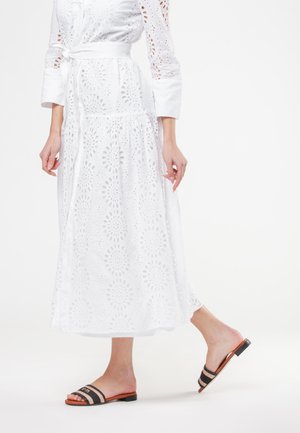 Donna che indossa un vestito bianco a maniche lunghe in broderie anglaise e sandali neri a righe con punta aperta e suola piatta, che cammina su uno sfondo bianco semplice.