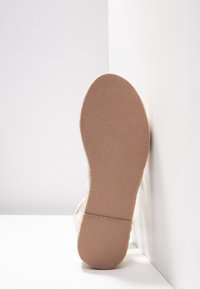 Semelle d'une chaussure, beige clair avec un motif de grip texturé, présentant un bord lisse et une forme incurvée, appuyée contre une surface blanche.